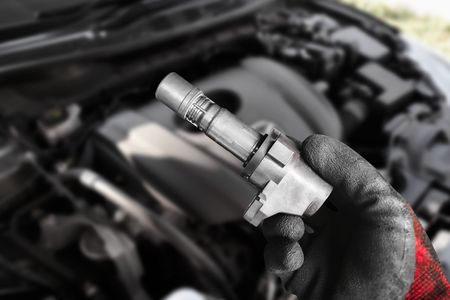 Hand holding a car ignition coil in front of an engine bay. Black gloves and red plaid sleeve visible.