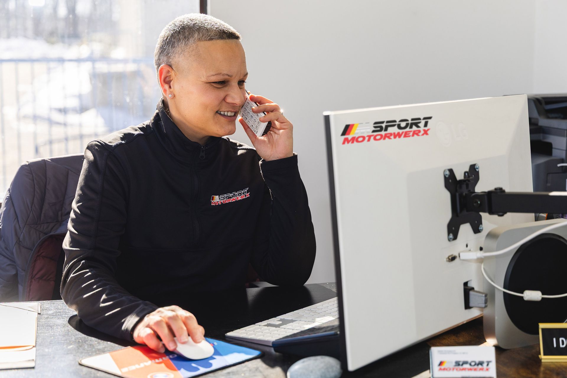 Woman in black jacket on the phone, working at a computer with M Sport Motorwerks logo.