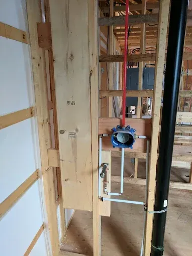 A shower valve with plumbing connections mounted between wooden wall studs at a construction site.