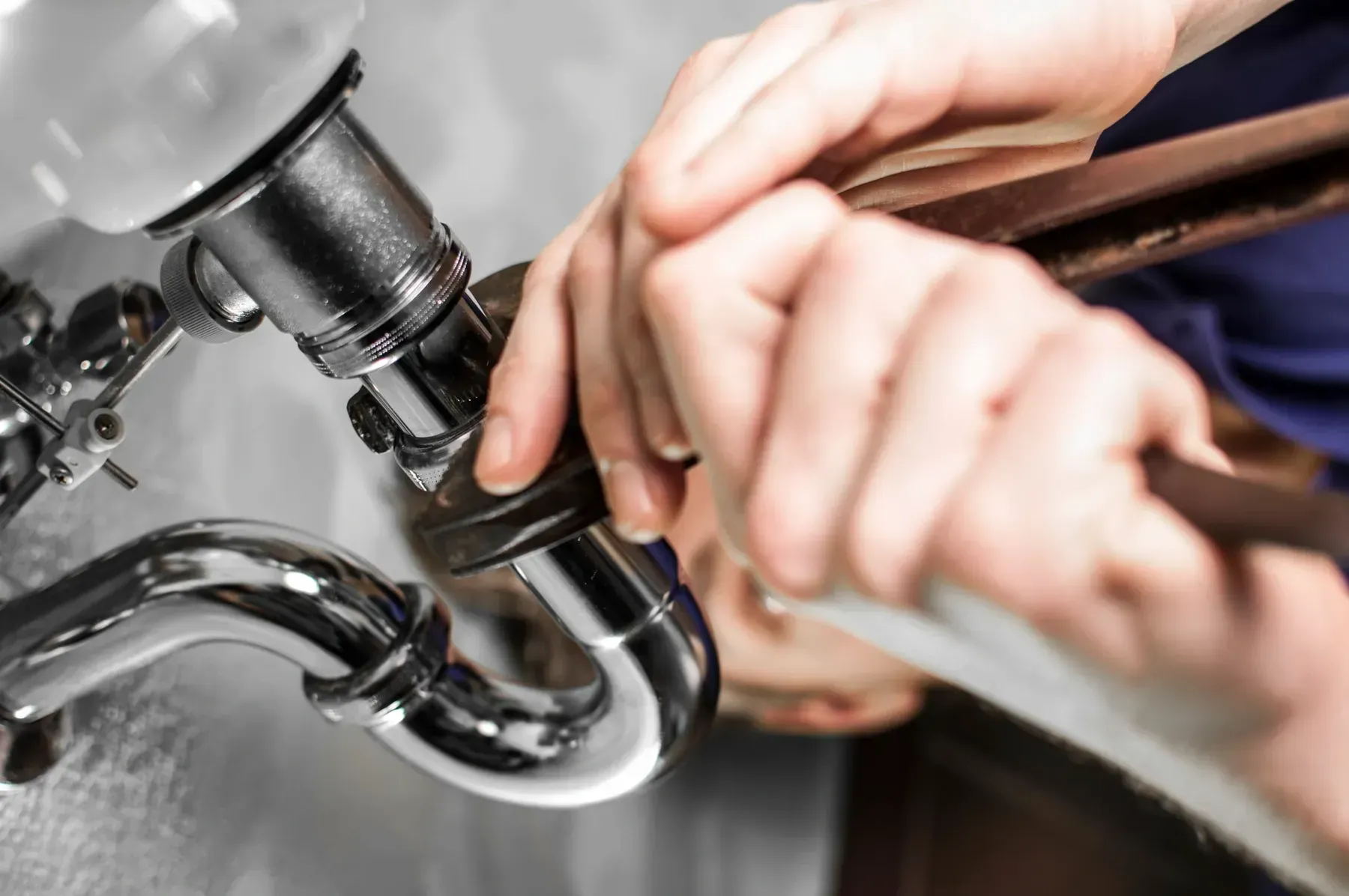 Close-up of hands using a metal wrench to tighten or loosen the chrome P-trap pipe under a sink.