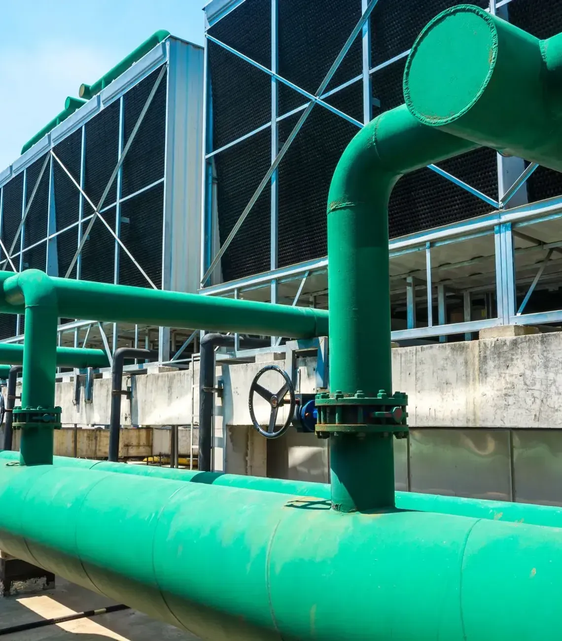 Large green industrial pipes with valves sit in the foreground of a row of outdoor cooling towers under a blue sky.