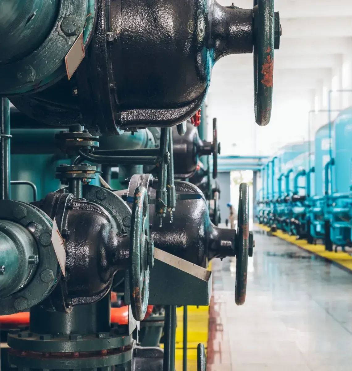 Dark industrial valves and piping in the foreground, with a row of blue water treatment tanks in an industrial facility.