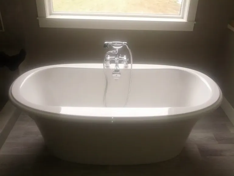 A white, oval, freestanding bathtub with a chrome faucet and handheld sprayer, centered in a bathroom before a window.