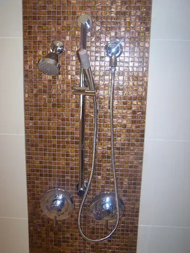 A shower wall with bronze-toned mosaic tiles, featuring a fixed showerhead, a hand-held sprayer on a rail, and two knobs.