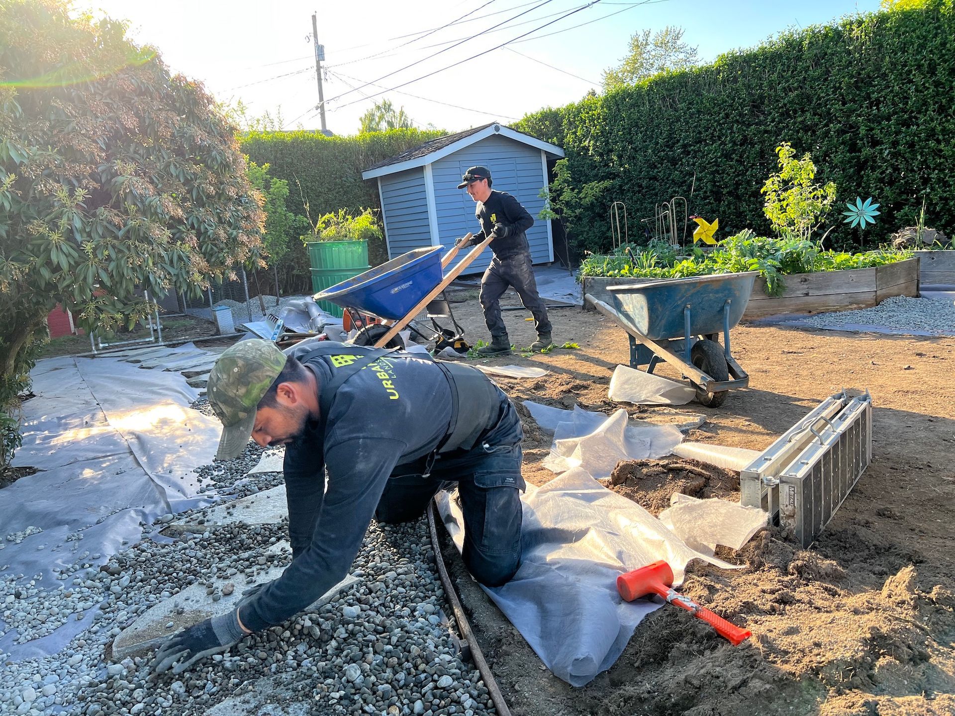 A man is kneeling down in the dirt while another man pushes a wheelbarrow.