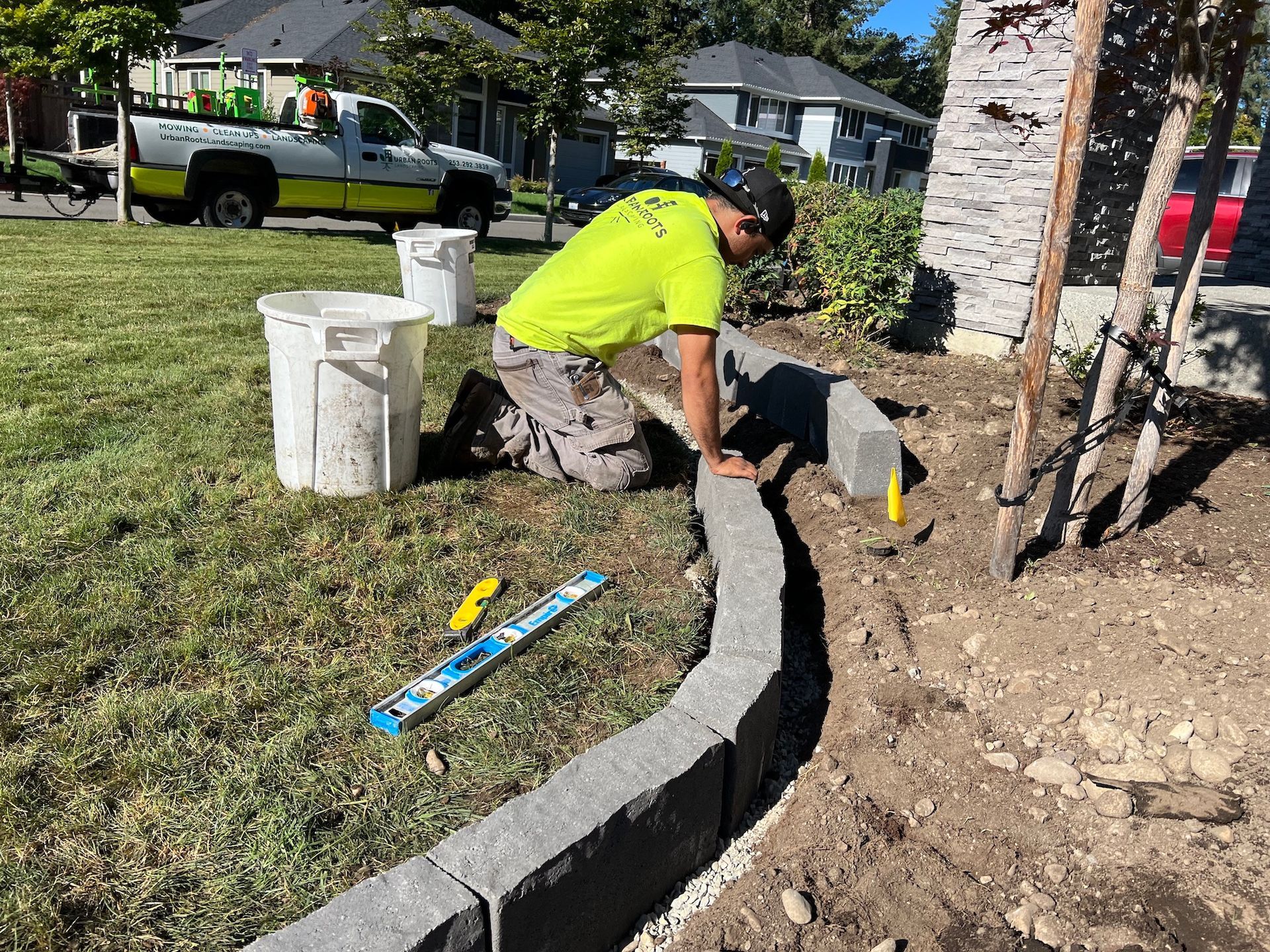 A man is kneeling down in the dirt while working on a concrete curb.