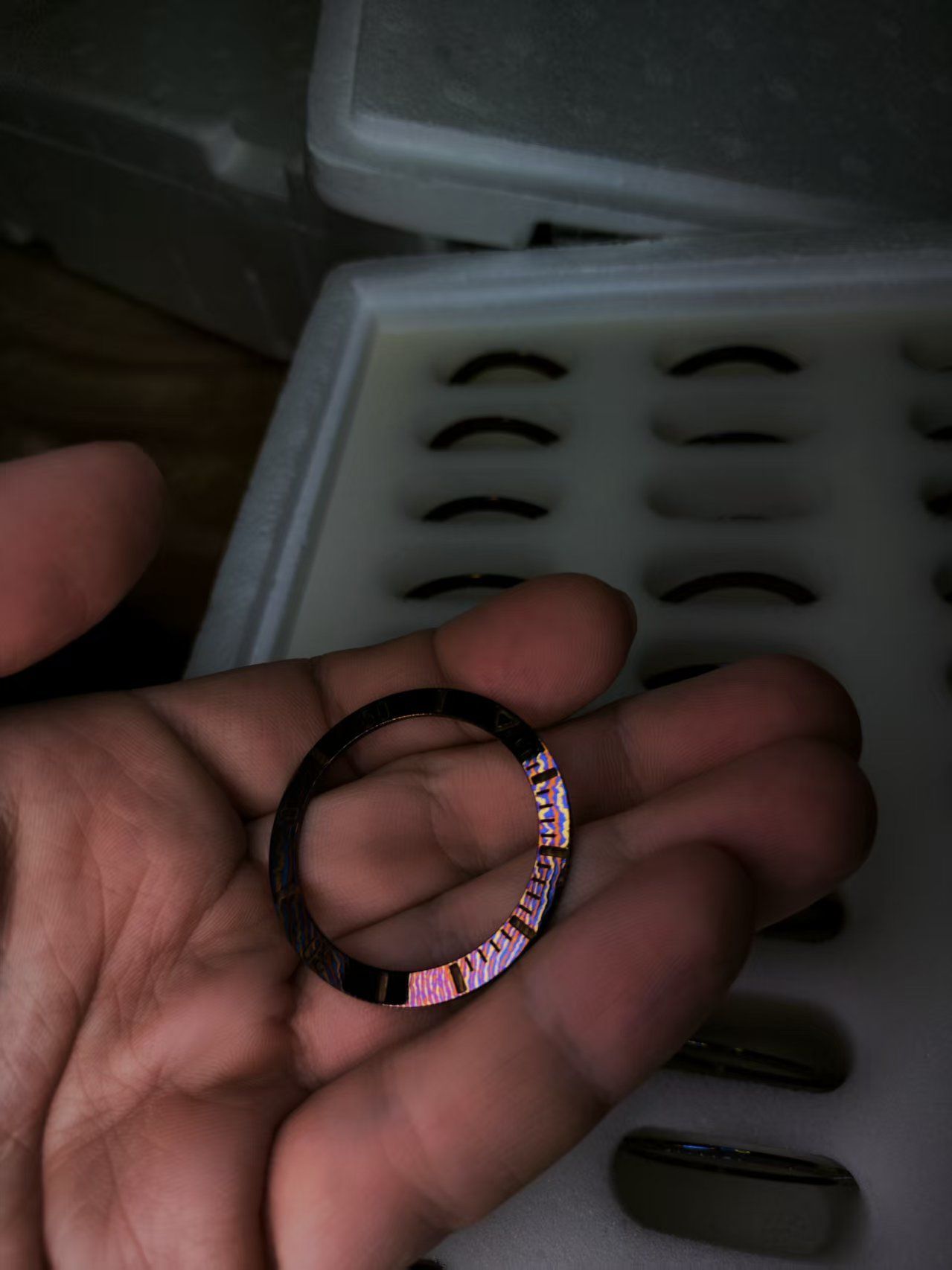 Hand holding a dark ring with etched text, in front of a tray with small curved items.