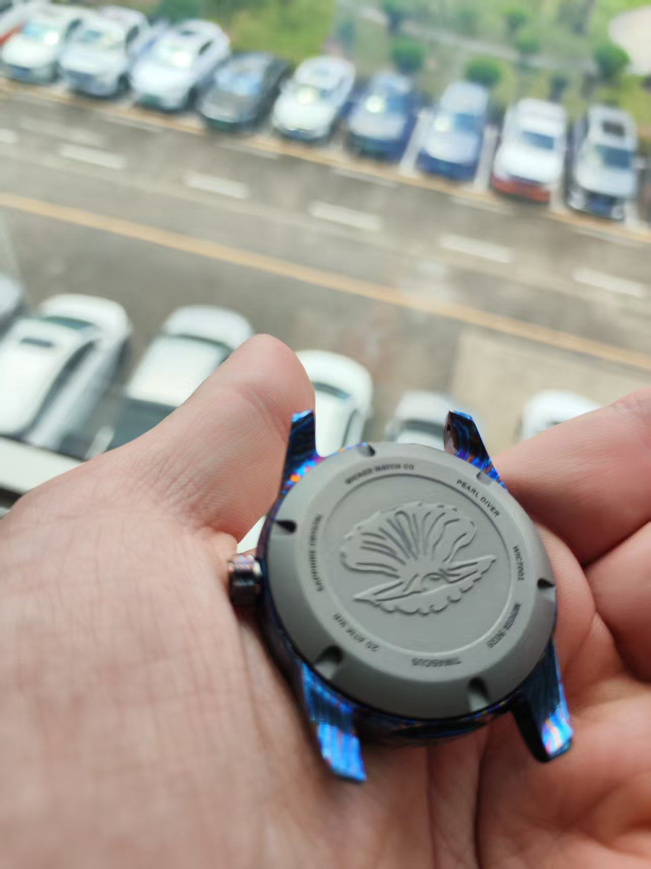 Person holding a wristwatch. The watch back is gray with a flower design, blue edges. Cars in the background.