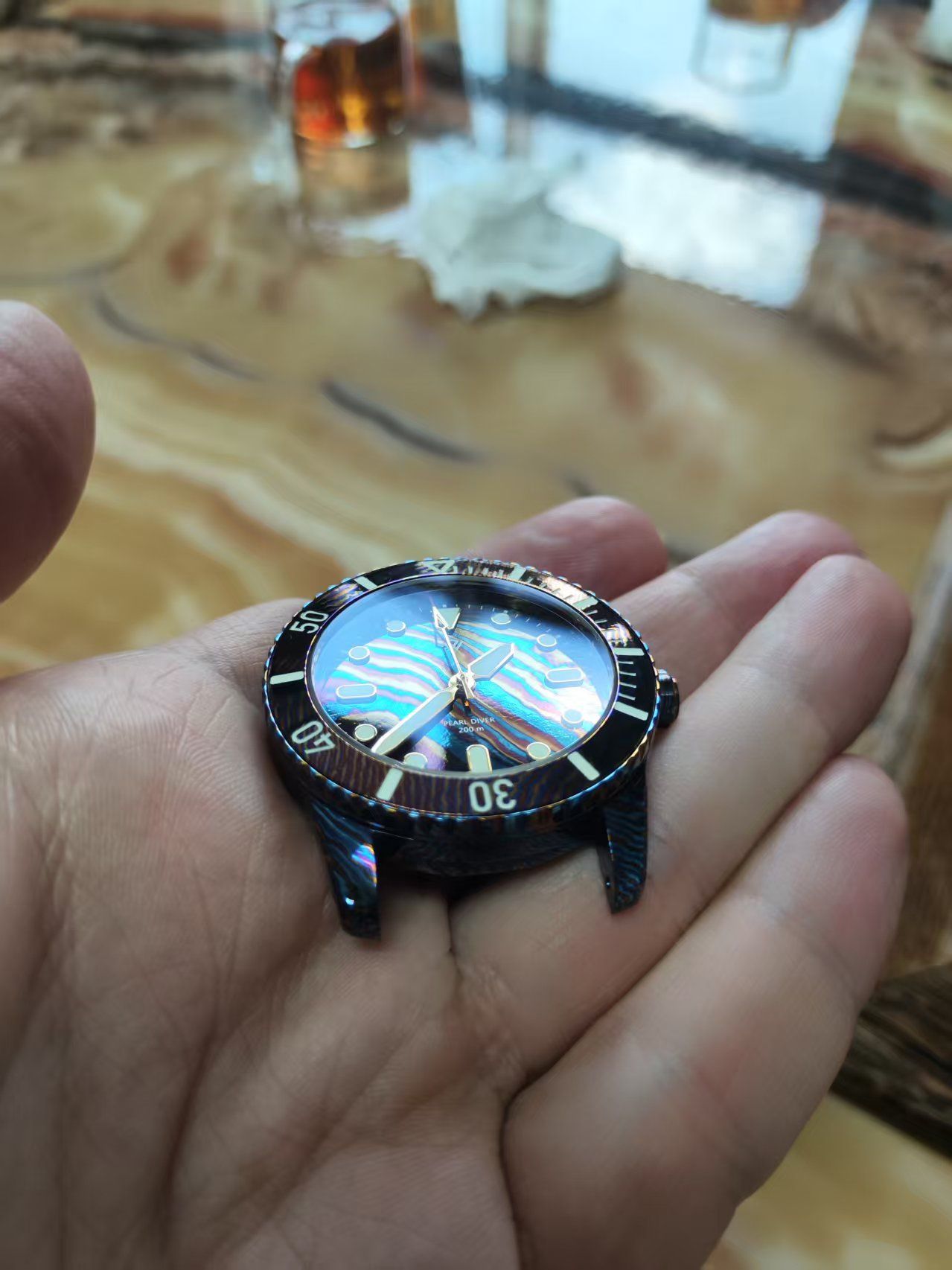 A wristwatch face with iridescent blue dial sits in an open hand. The watch has a black bezel and lugs.