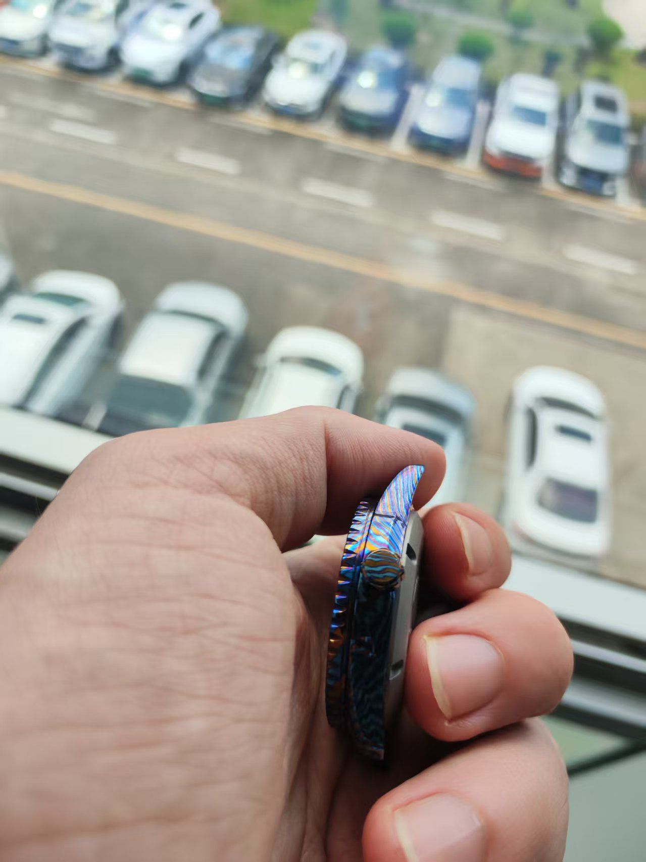 Hand holding a small, colorful, metal item with a blurred background of parked cars.