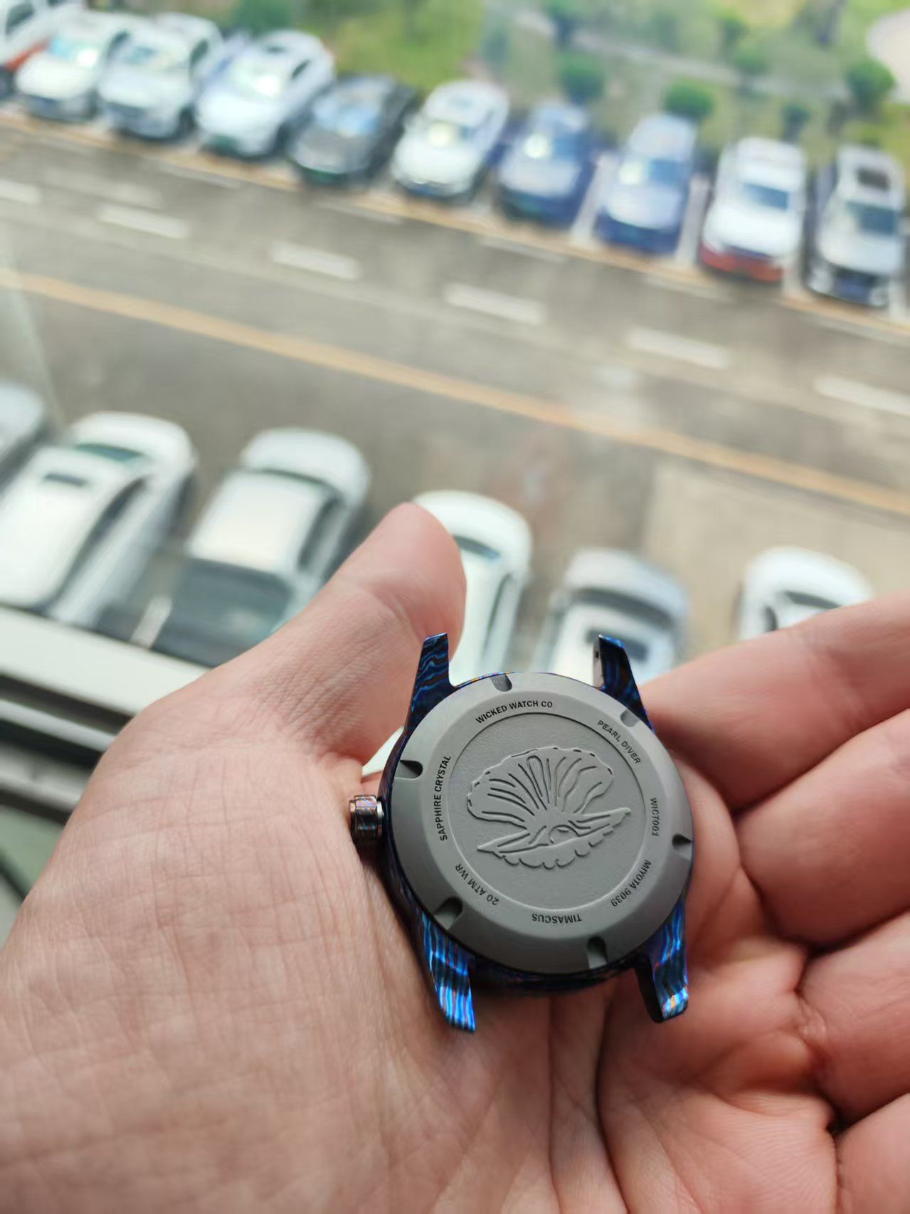 A person holding a watch with a blue frame, the watch back displaying design. Cars in background.