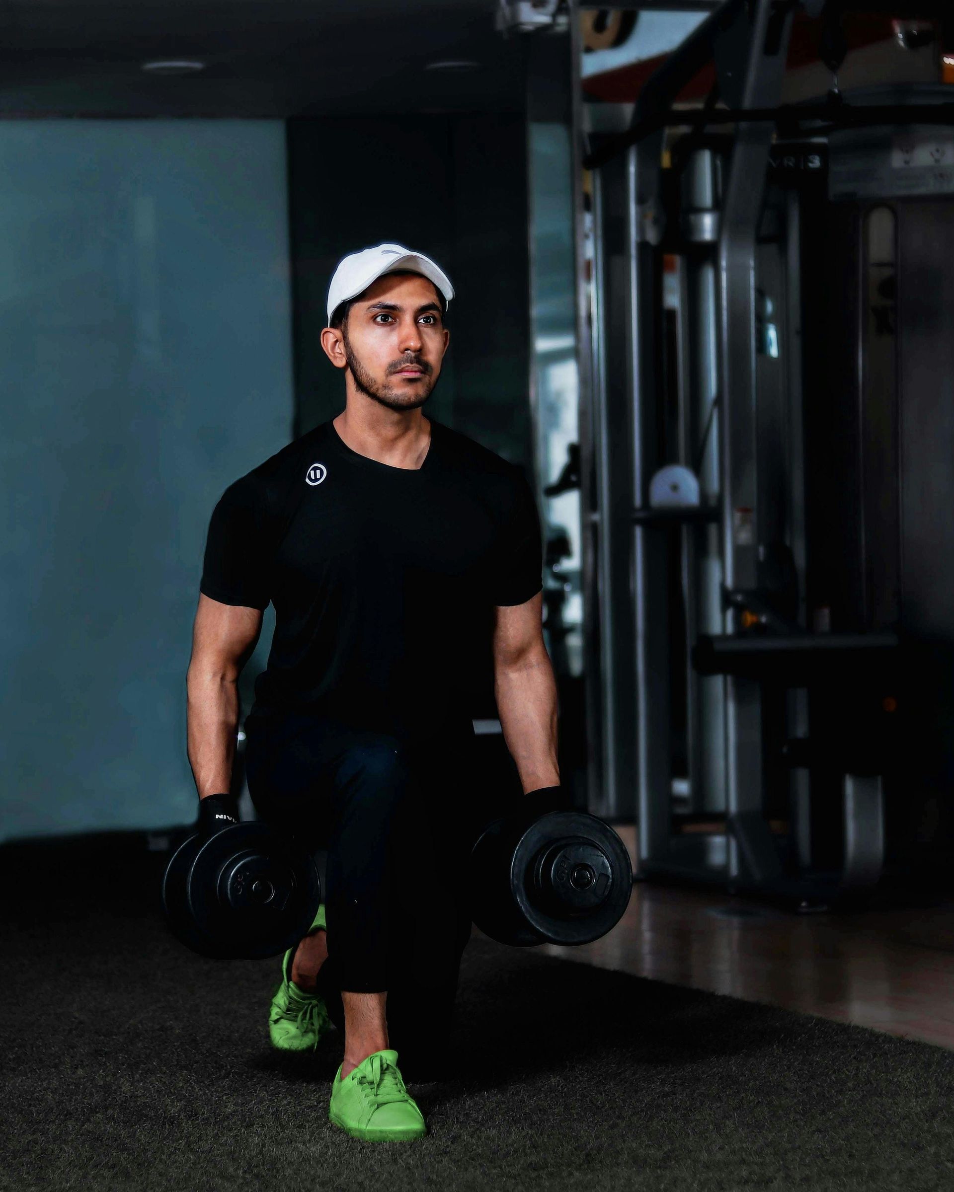 A person wearing a white cap performs a dumbbell lunge in a gym, wearing a black shirt, black pants, and neon green shoes.