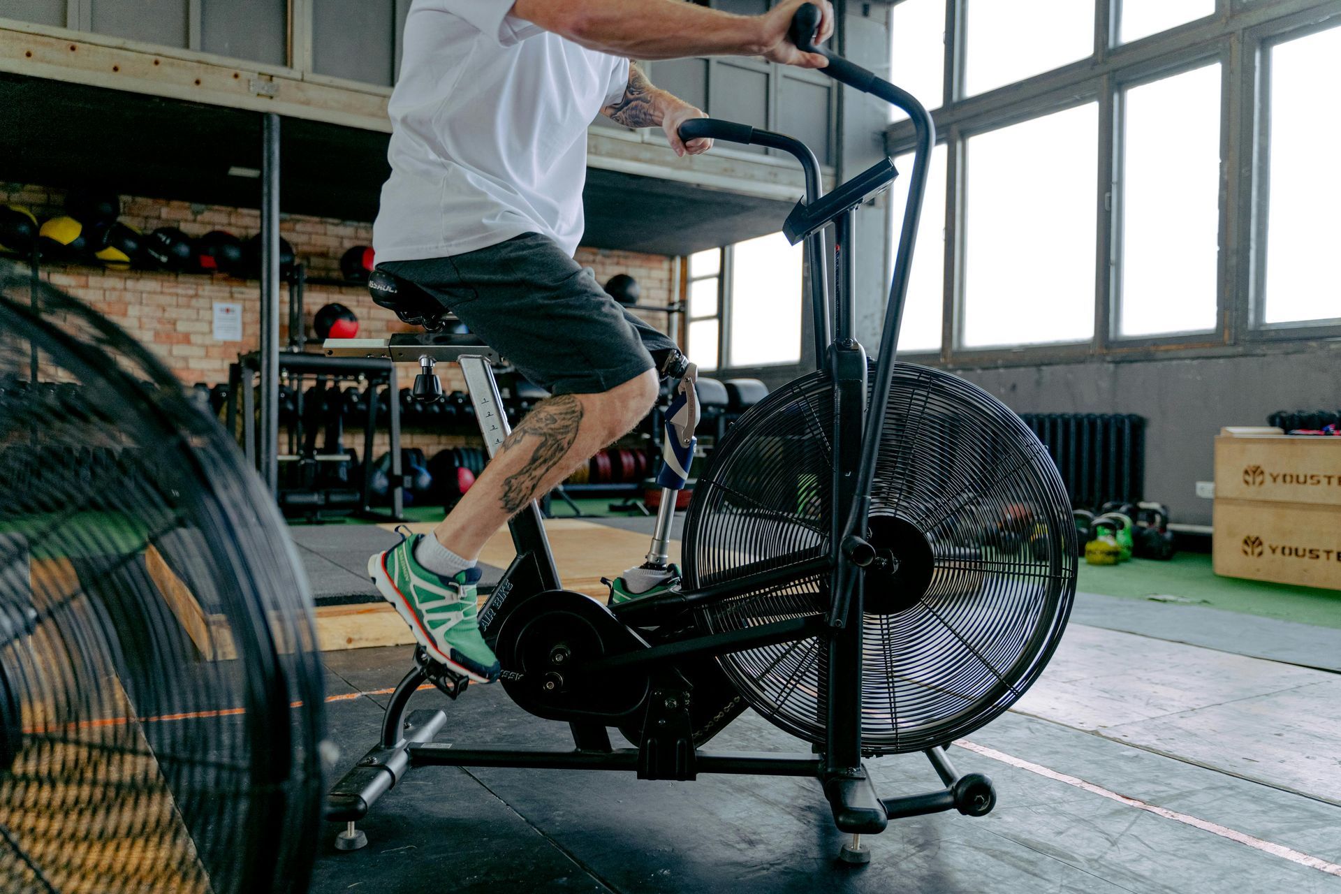 A person pedals an air bike in a gym, wearing a white shirt, dark shorts, and green athletic shoes.