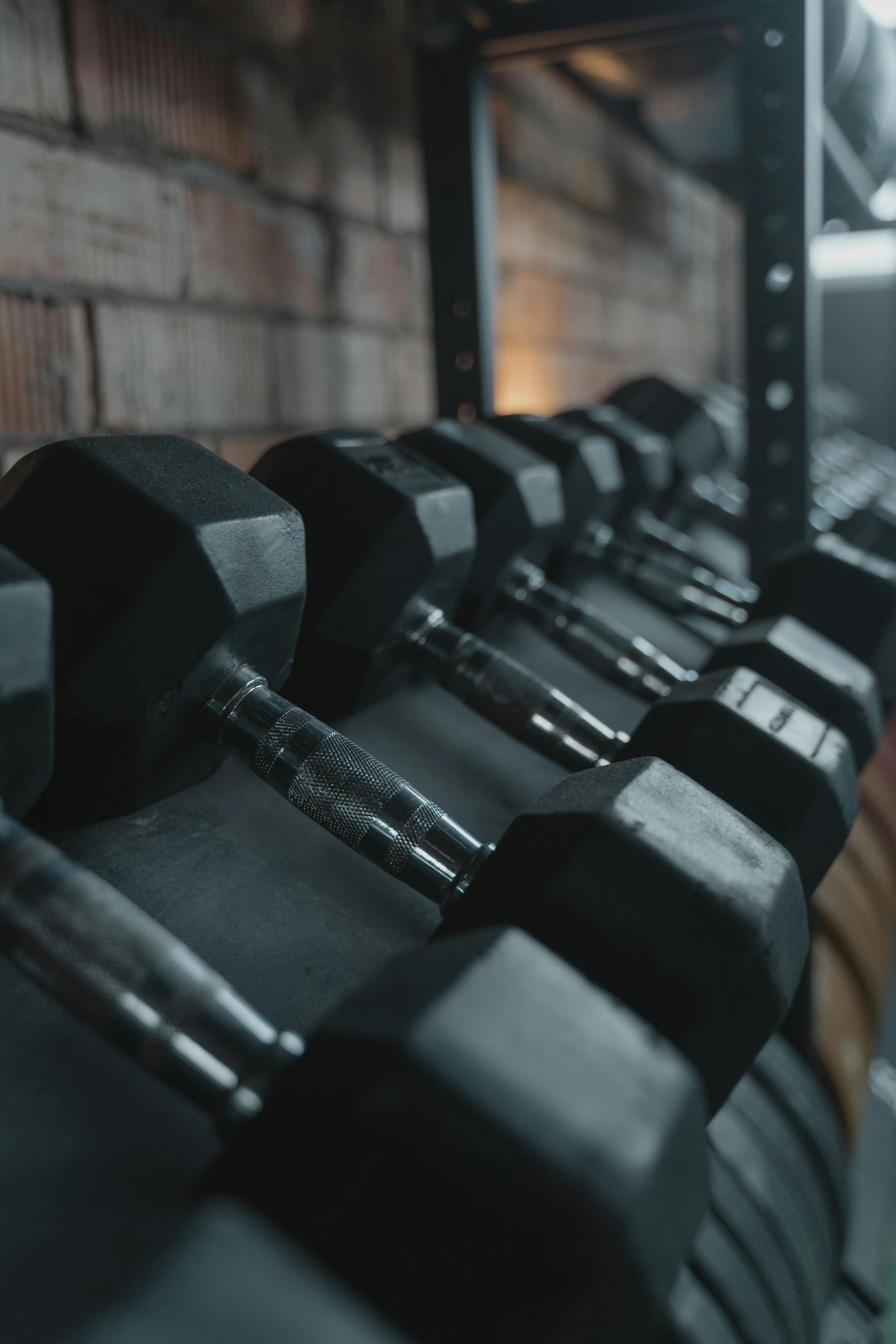 A rack of black, hexagonal dumbbells lined up in a gym with a brick wall background.