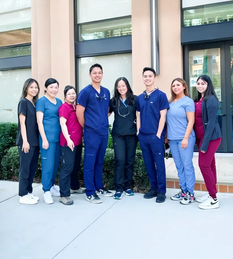 Dental team stands outside a building. They wear scrubs in various colors. Smiling, posing for a photo.