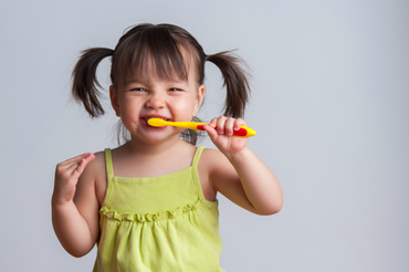 Young girl with pigtails, wearing a green top, brushing teeth with a yellow toothbrush, smiling.