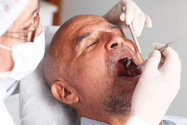 Man at dentist, mouth open, being examined with tools. Dentist in mask and gloves.