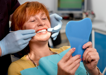 Woman in dental chair examining her teeth with a mirror while dentist checks with a tool.