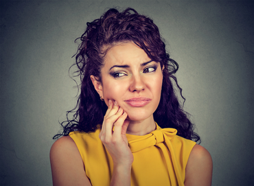 Woman with curly hair, in a yellow top, touching her jaw with a pained expression; gray background.
