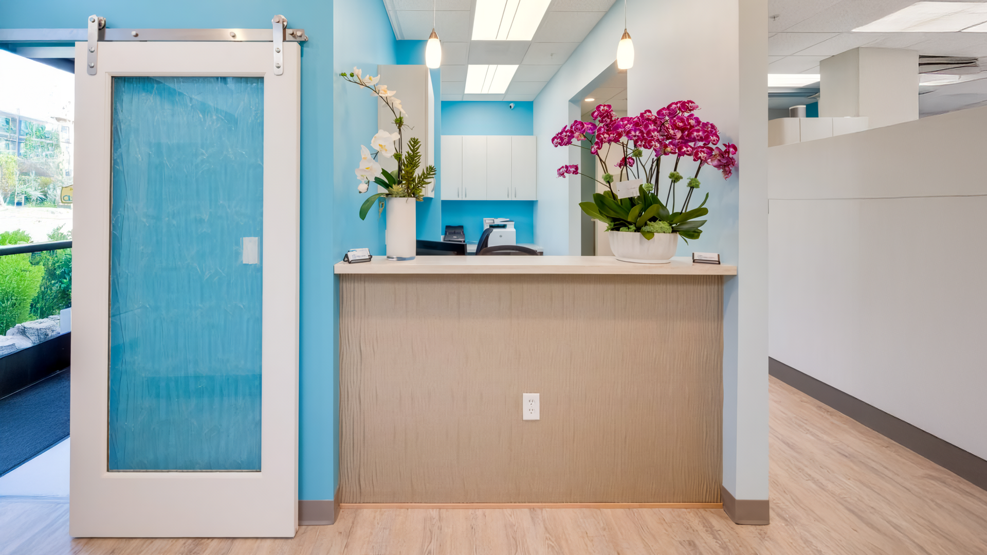 Reception area with a light wood desk, turquoise wall, orchids, and a sliding door.
