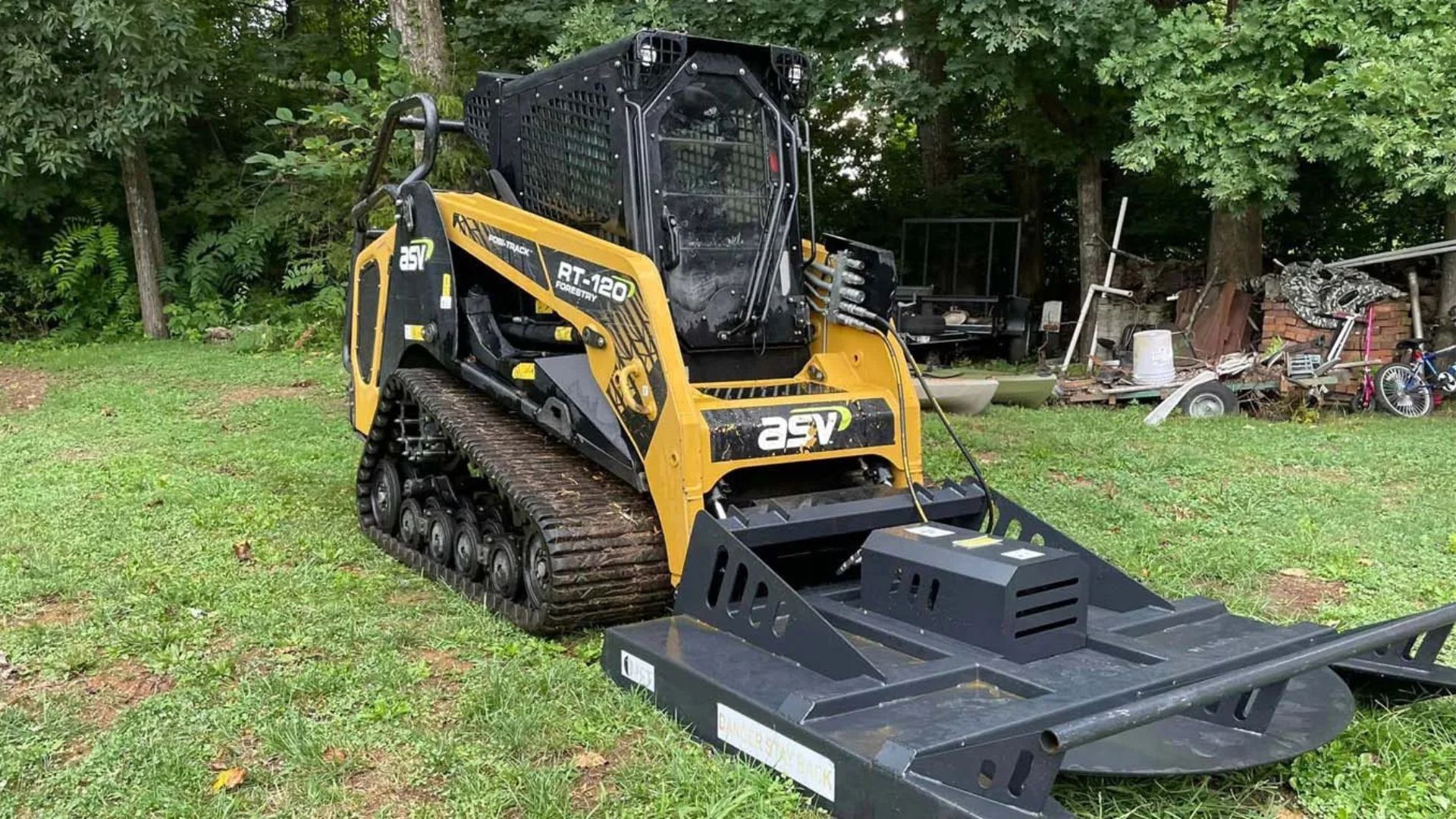 Yellow and black track skid steer with brush cutter attachment on a grassy lawn.