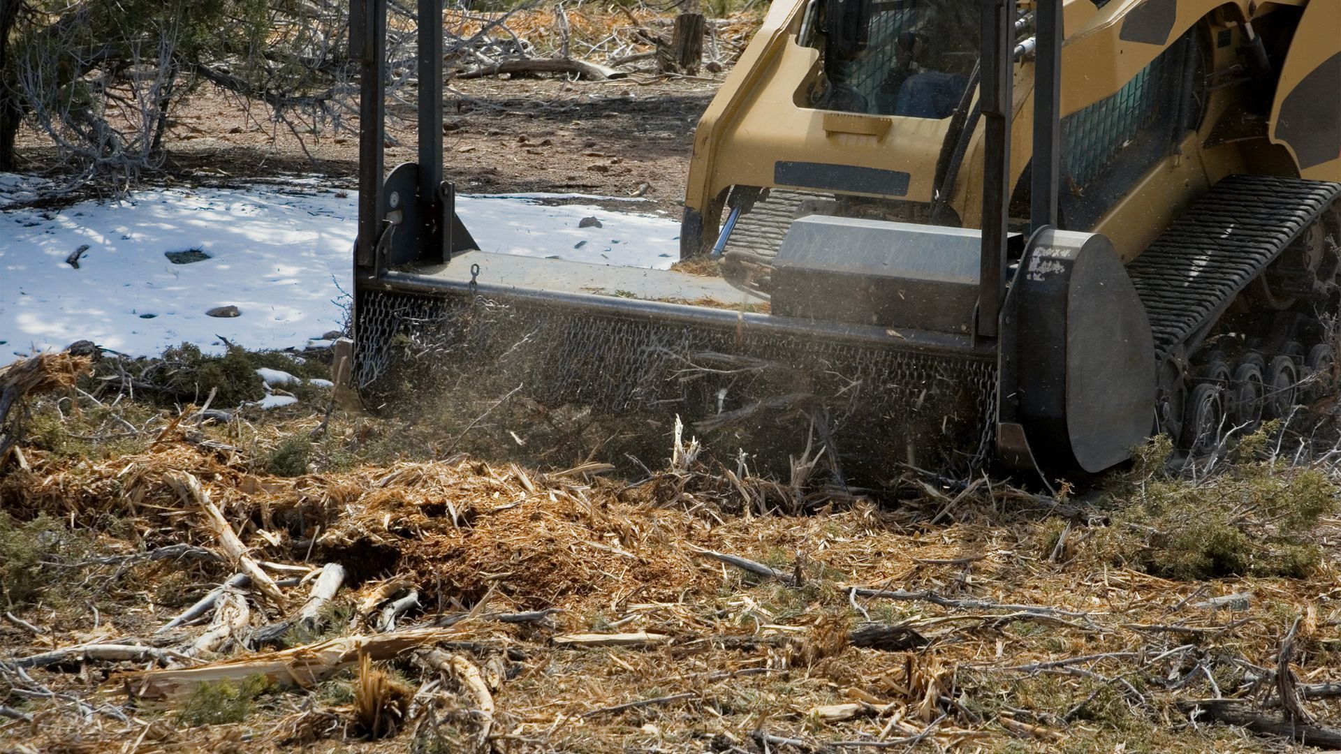A skid steer mulching brush, creating wood chips. Brown machine, debris in snowy landscape.