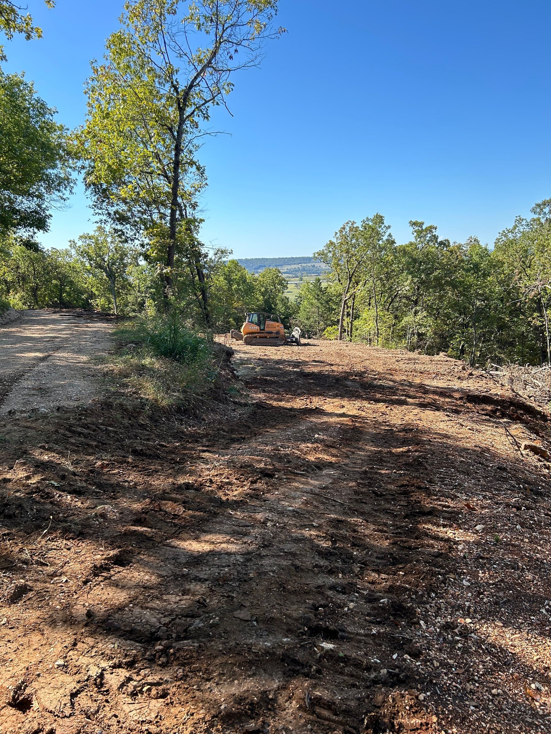 Dirt path through a wooded area; small construction vehicle in the distance, bright blue sky.