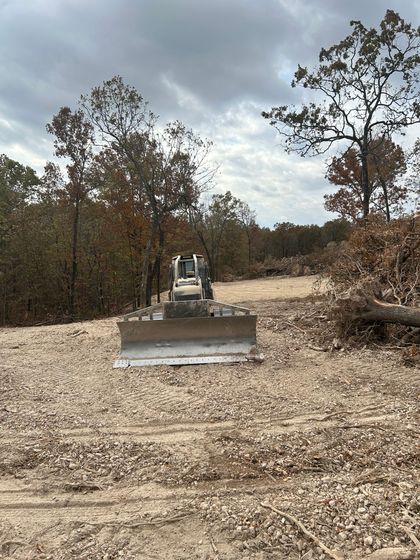Bulldozer on a cleared, gravelly field surrounded by trees under a cloudy sky.