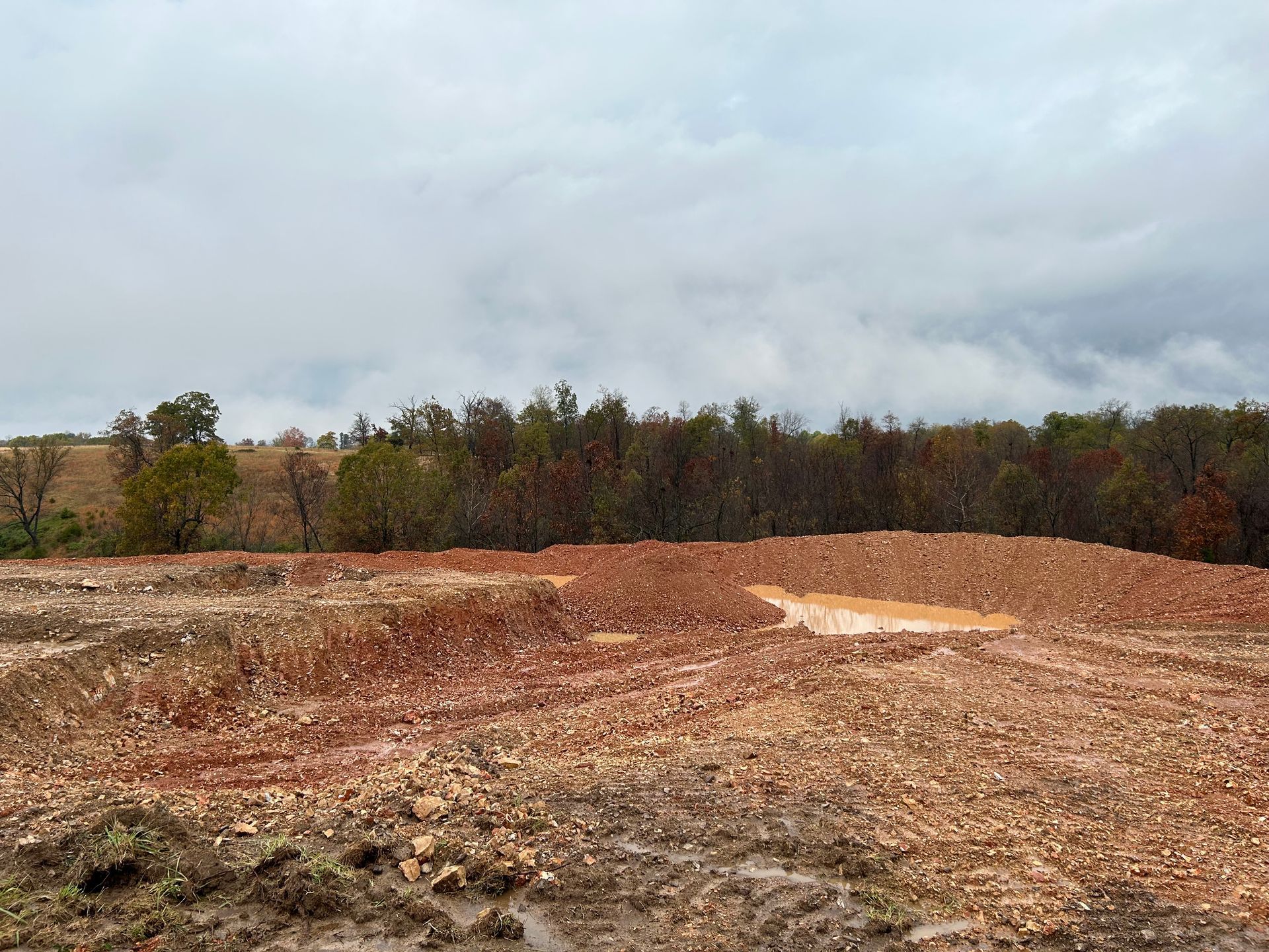 Muddy, reddish-brown earth and piles in a field. A line of trees is in the background under a cloudy sky.