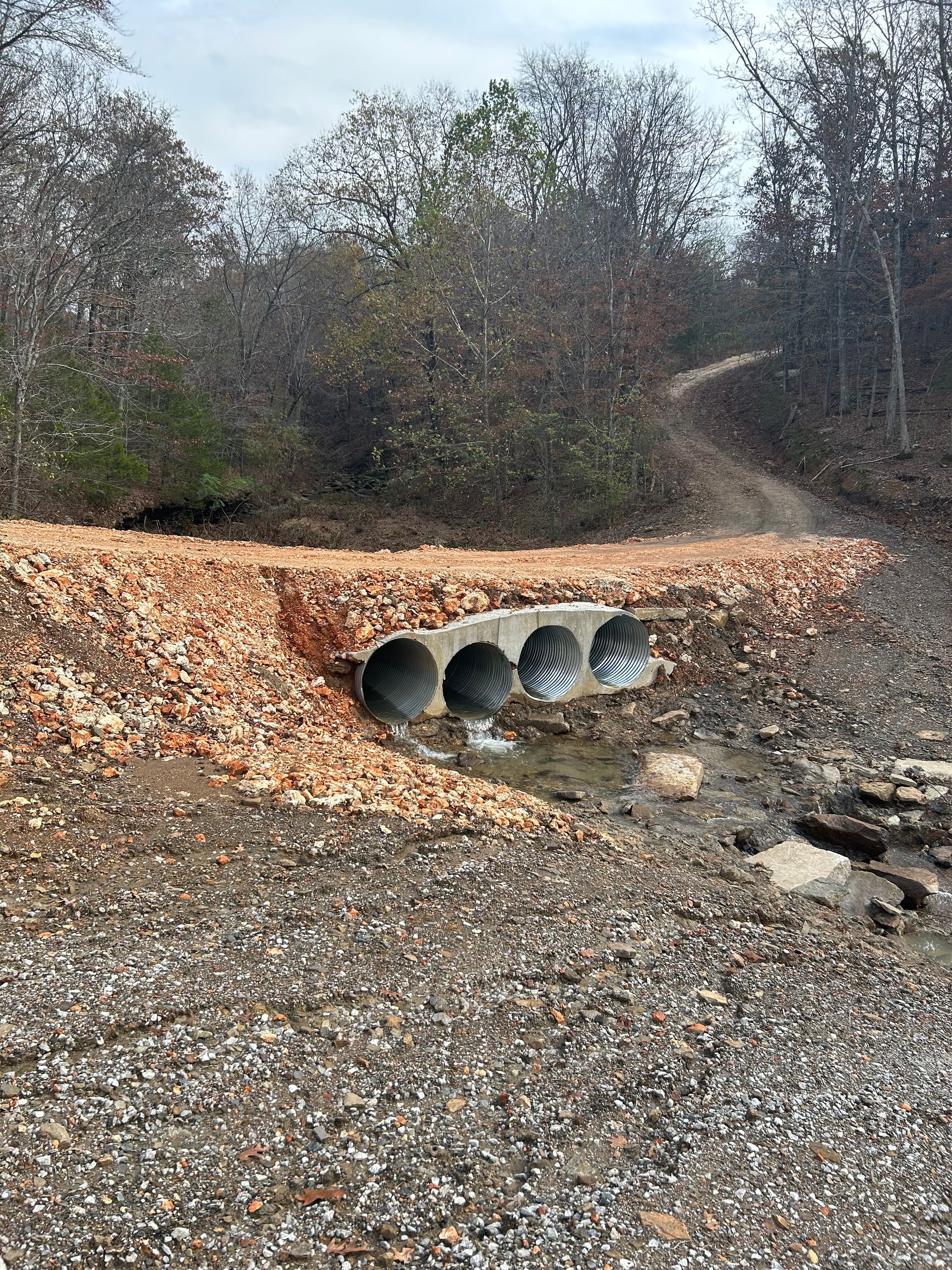 Concrete bridge over a small stream, part of a trail. Brown gravel and a treeline in the background.