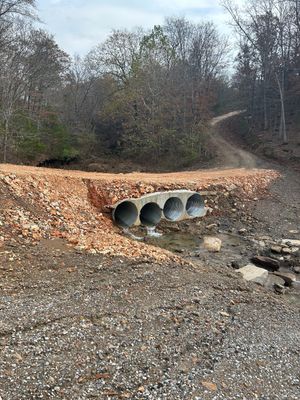 A gravel path crossing a creek through concrete culverts, lined with brown stone. Overcast day.