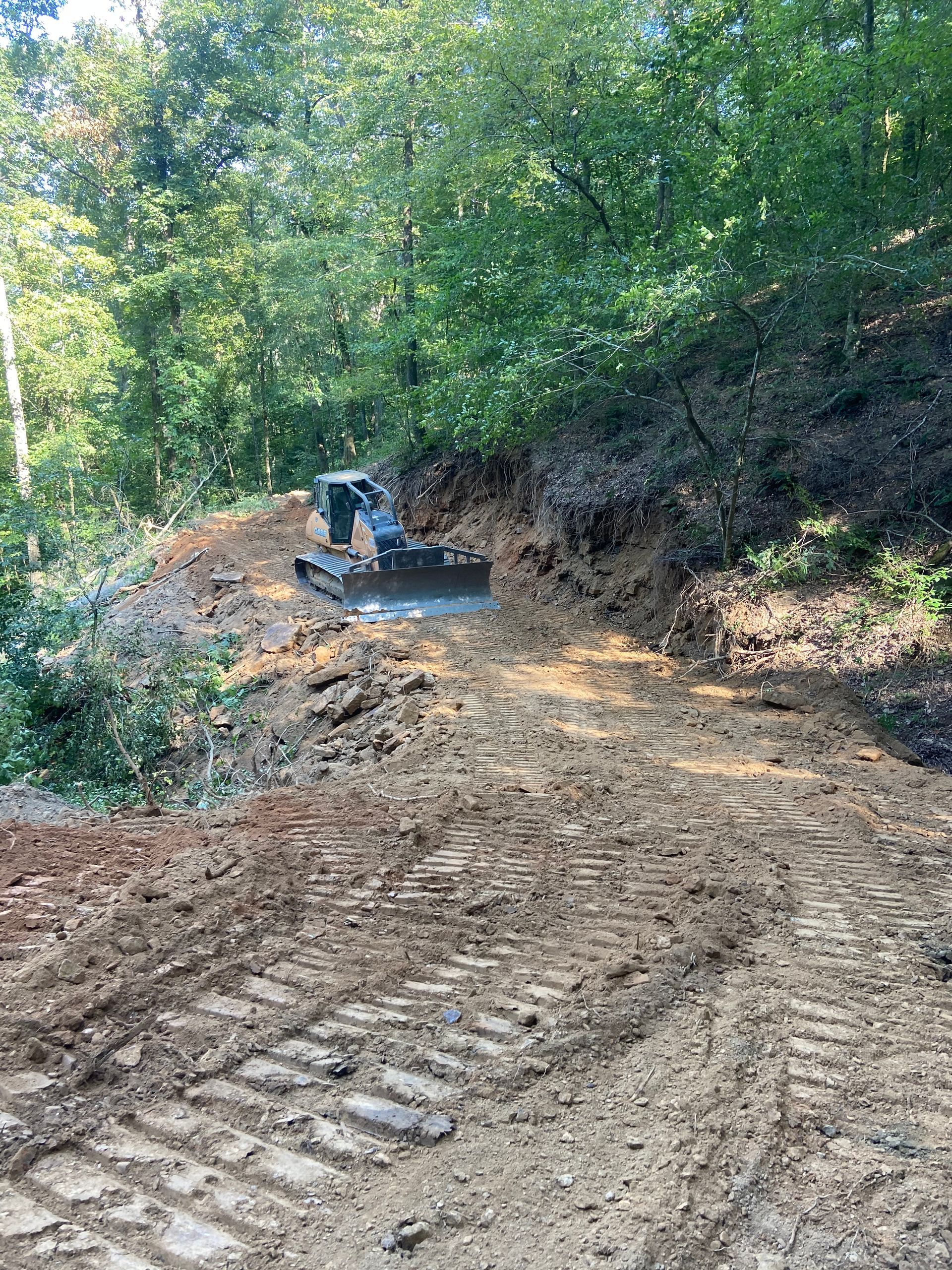 A tracked excavator working on a muddy dirt road in a wooded area.