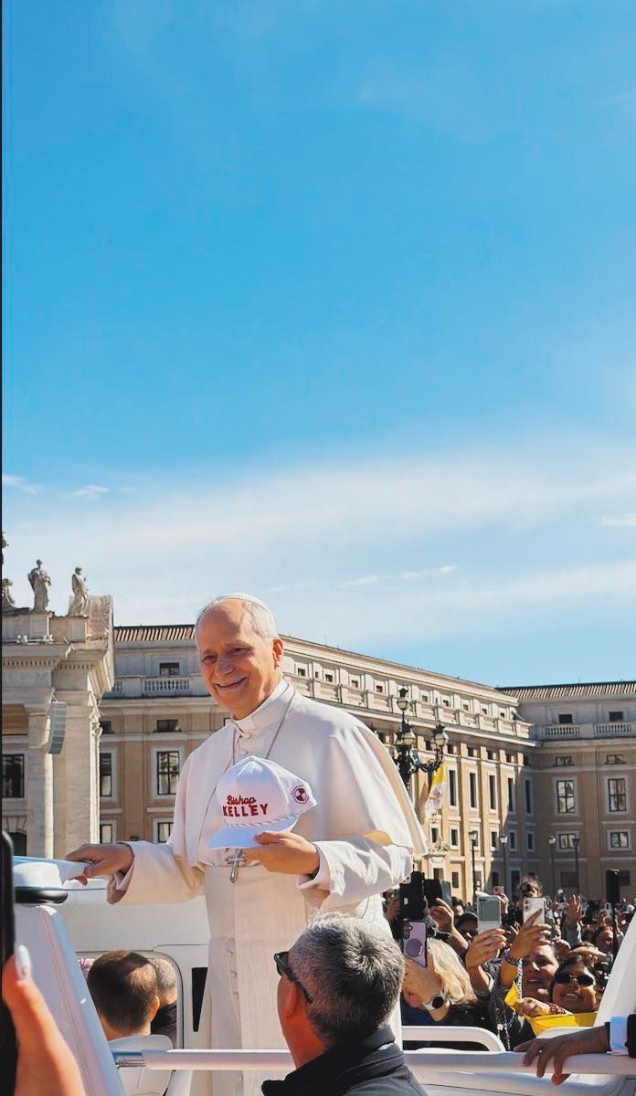 Pope Francis smiles and holds up a white baseball cap with 