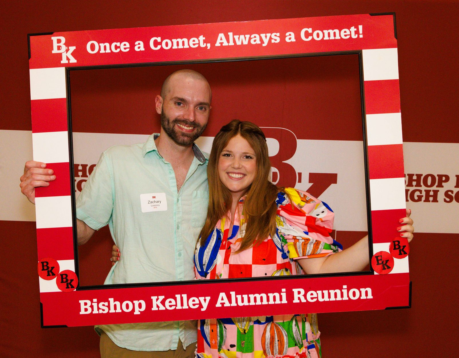 Two people smiling, posing behind a photo frame at a Bishop Kelley Alumni Reunion, red background.