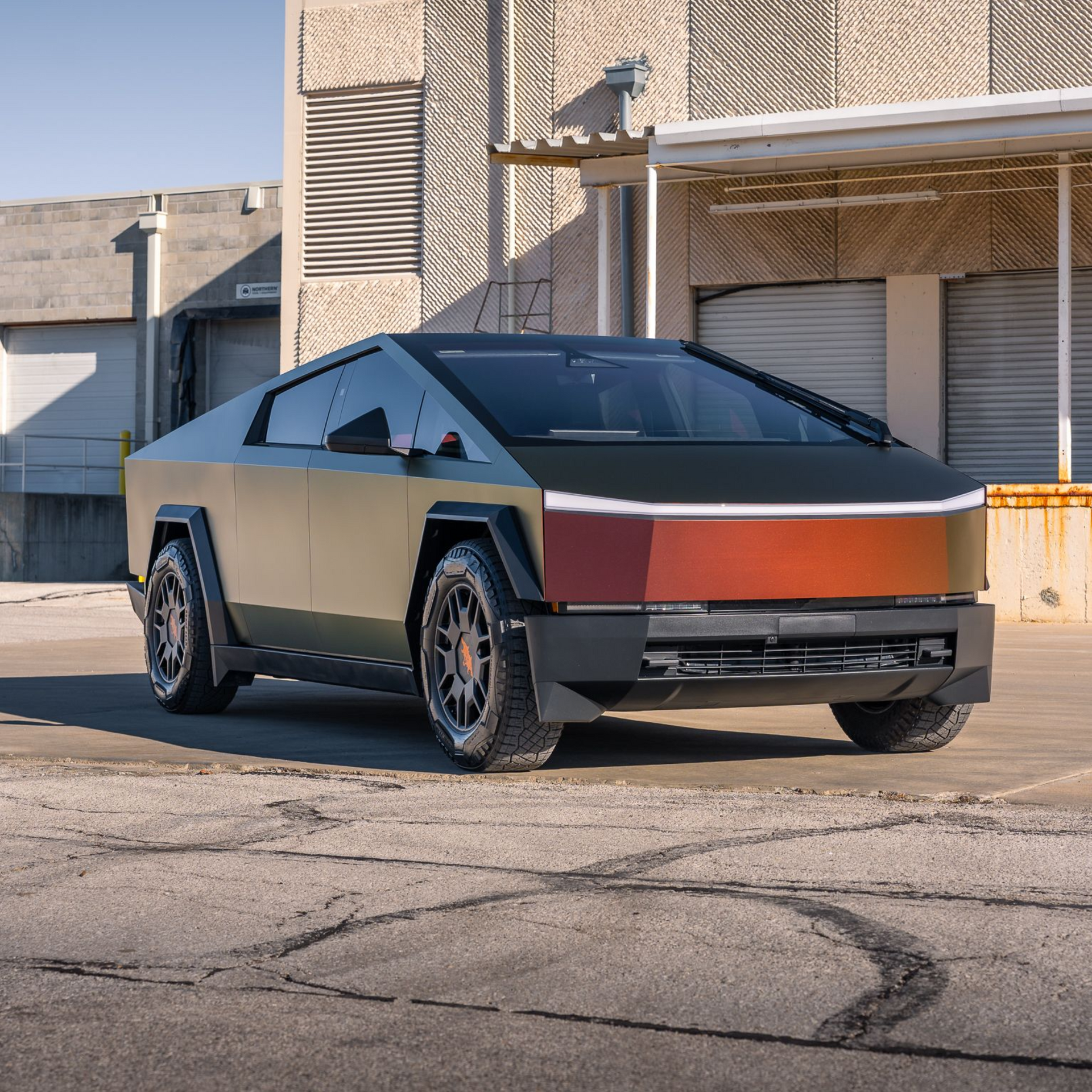 Tesla Cybertruck parked in an industrial alley, shown in matte silver, black, and orange panels.