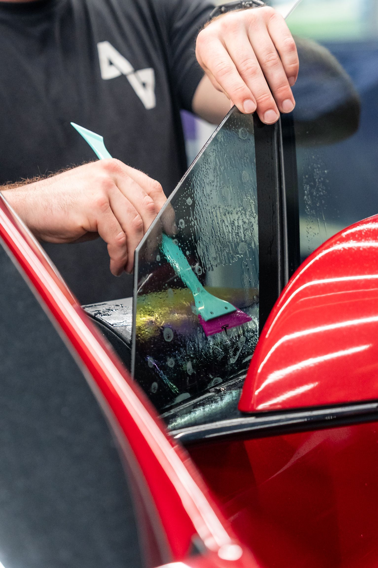 Hands cleaning a red car window with a squeegee during window tint installation.