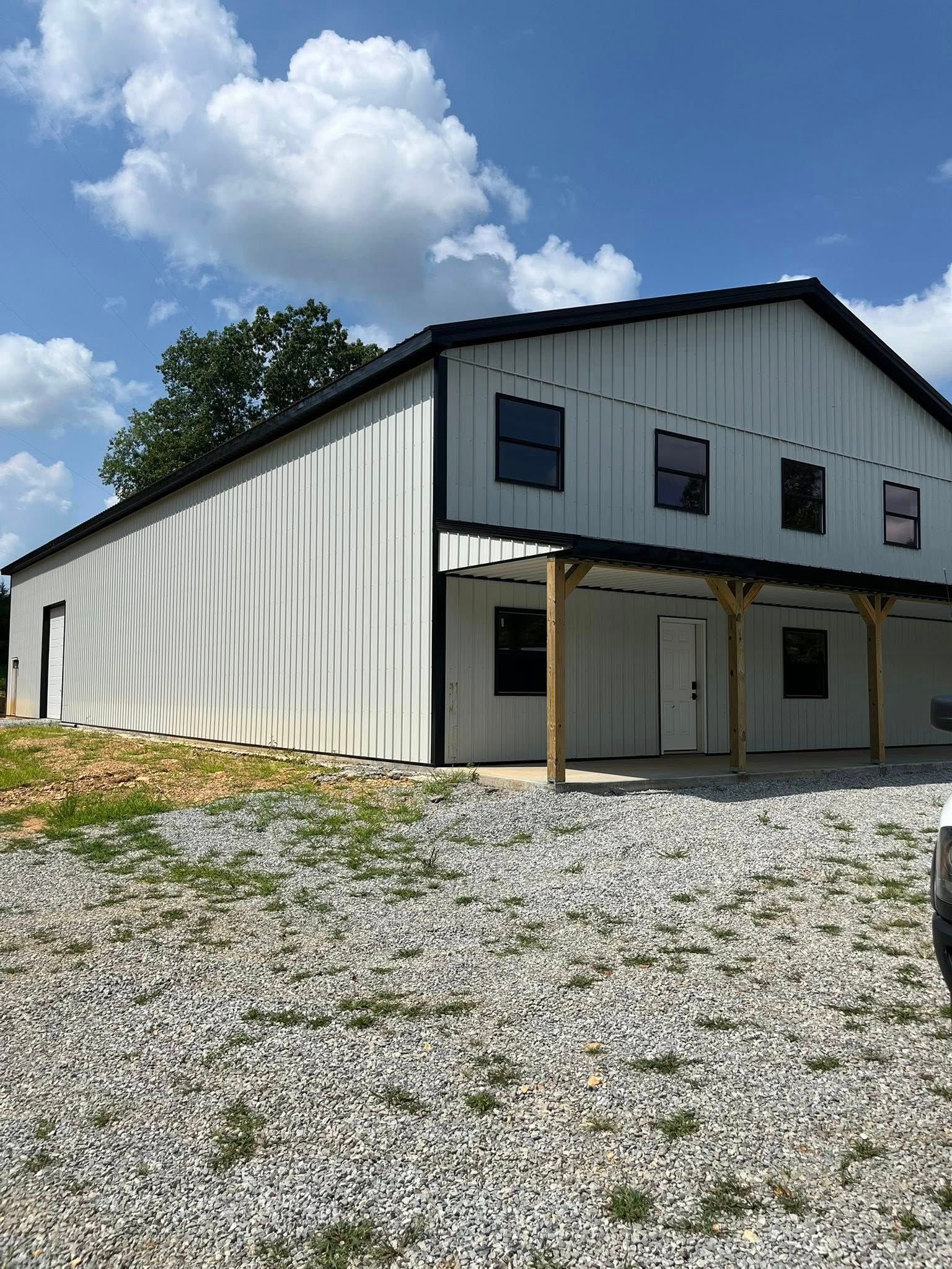 A white two-story metal building with a black roof and porch, situated on a gravel lot under a sunny, blue sky.