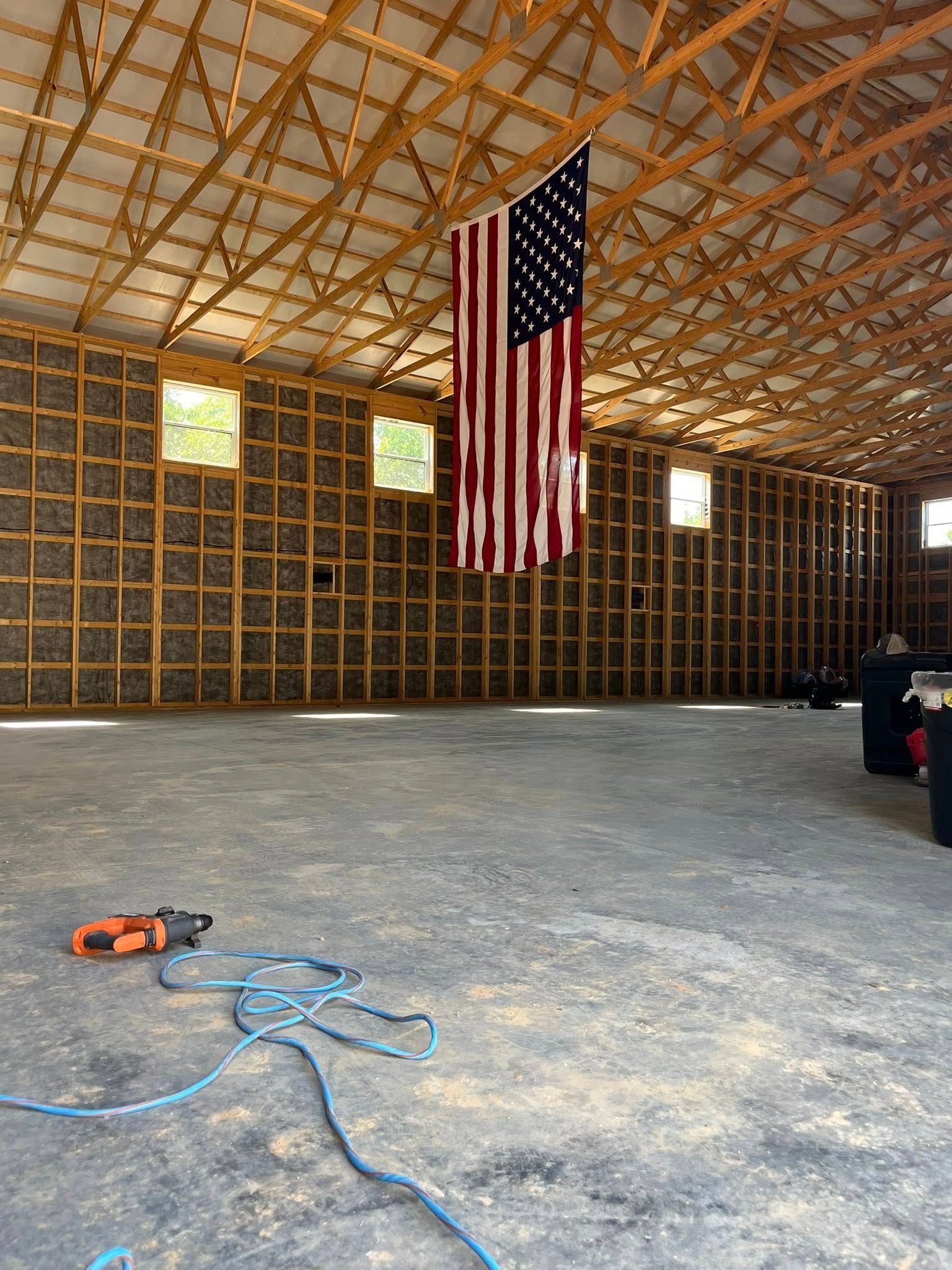 An American flag hangs from the rafters of a large, unfinished wooden building with a concrete floor and power tool.