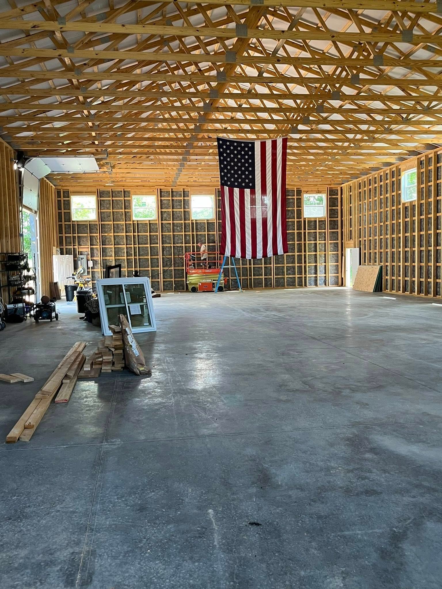 Large, unfinished barn interior with wooden studs, a concrete floor, and an American flag hanging from the ceiling.