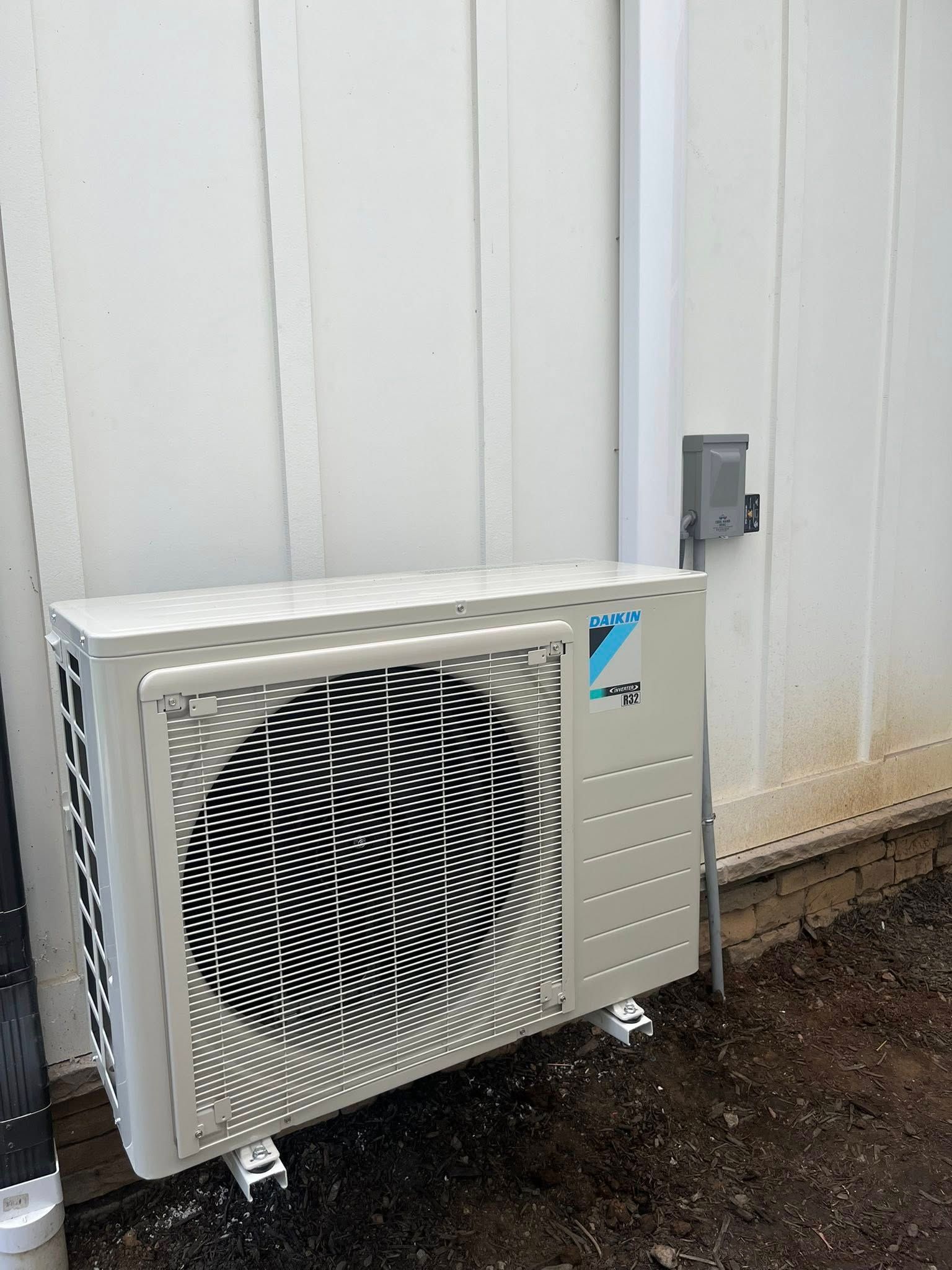 A beige Daikin outdoor HVAC unit mounted on a light wall next to an electrical disconnect box and a vertical white conduit.