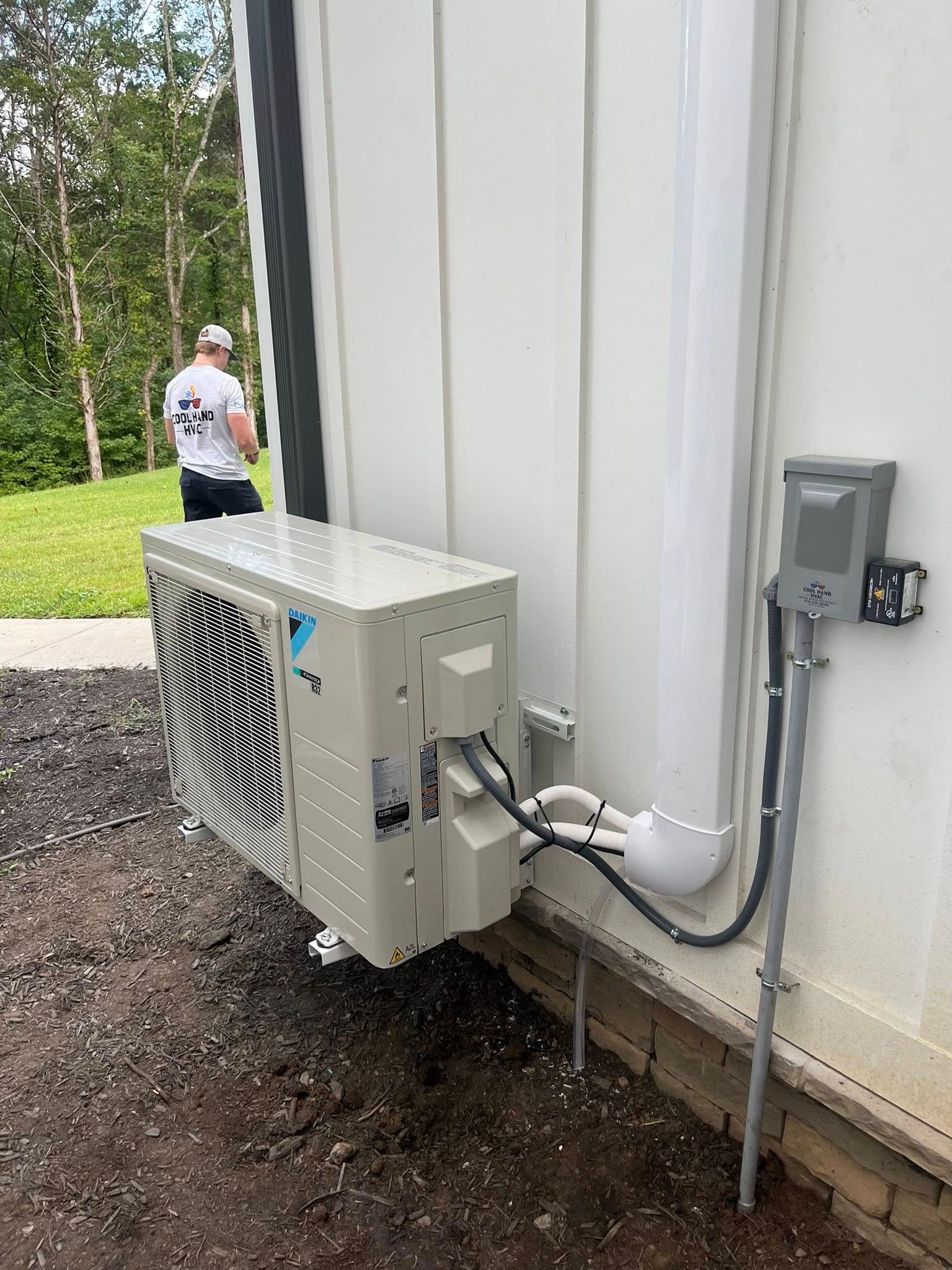 A technician stands near a beige Daikin outdoor HVAC unit mounted on a wall with electrical conduit and line covers.