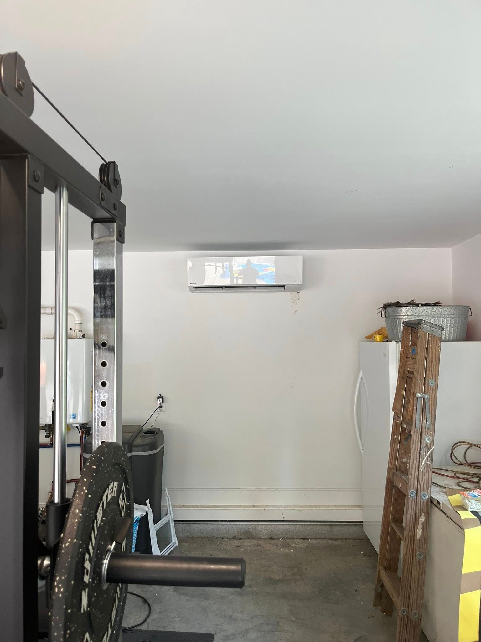 A wall-mounted ductless air conditioner unit installed in a garage with gym equipment and storage items.