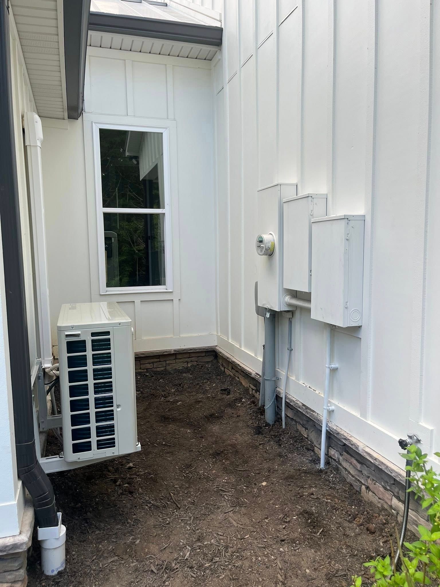A white exterior wall featuring an air conditioning unit and electrical boxes above a patch of dark, bare soil.