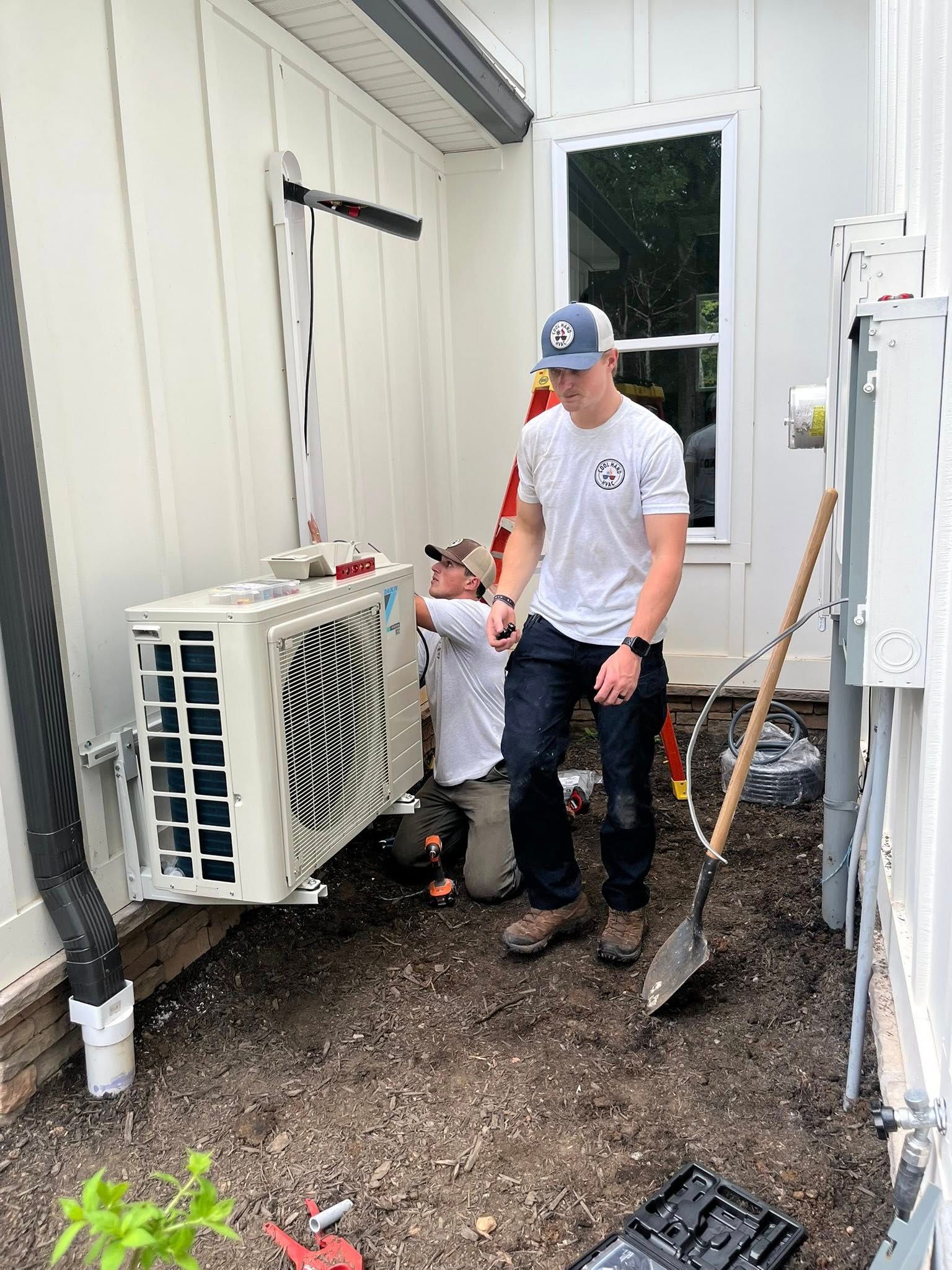 Two technicians installing an outdoor HVAC unit against the white siding of a building.