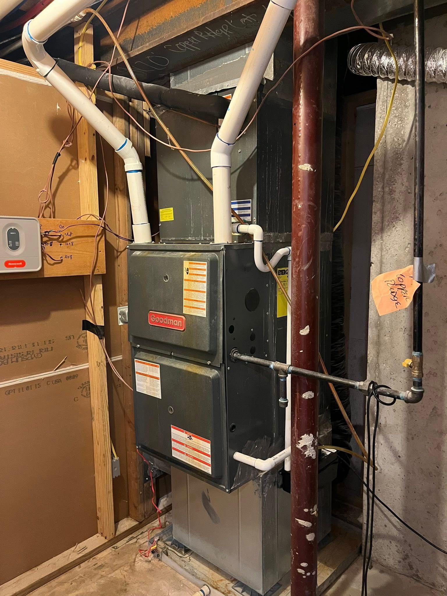 A grey residential furnace stands in a basement next to white PVC exhaust pipes, a metal support pole, and wiring.