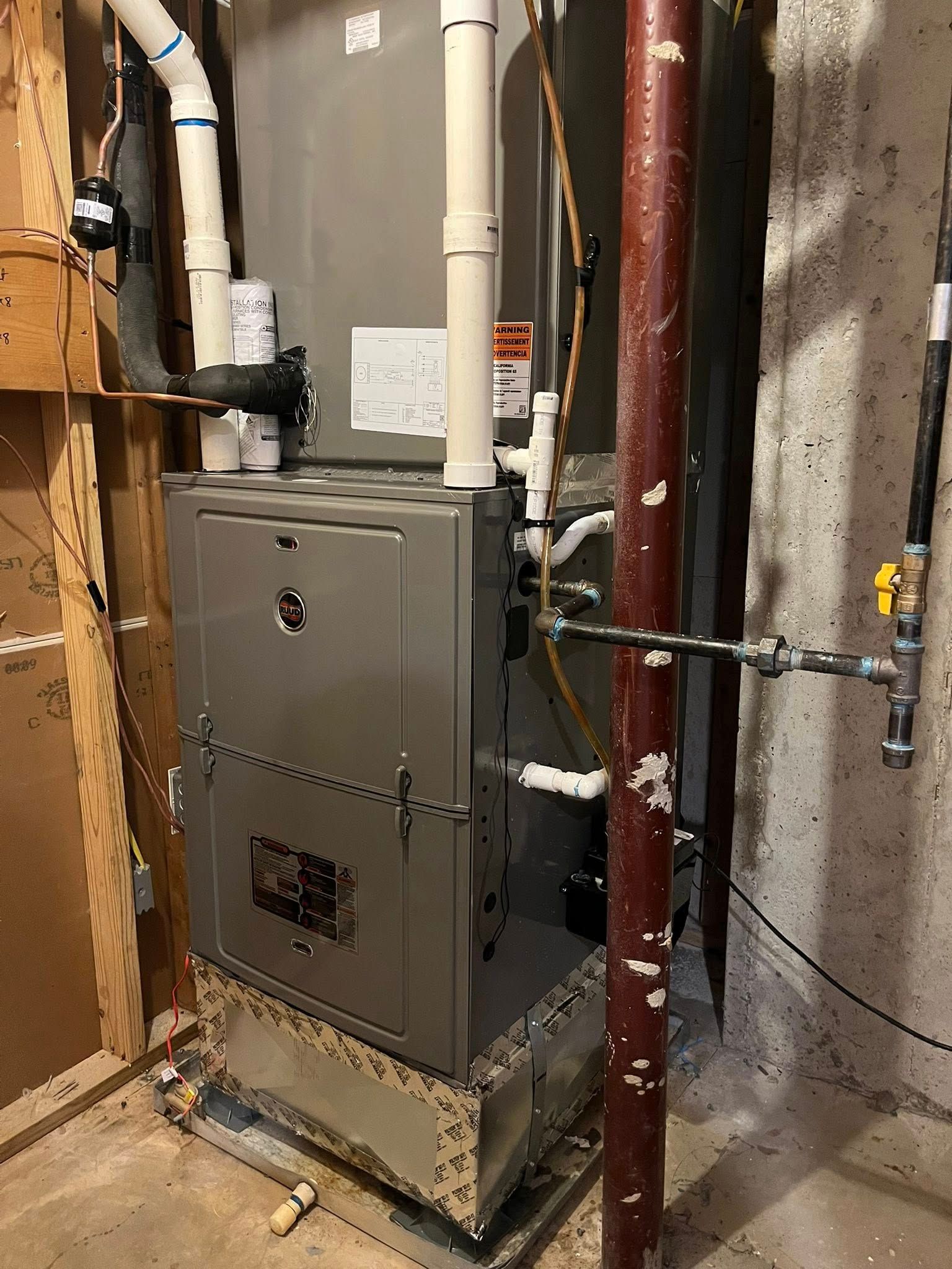 A grey furnace installed in a basement, featuring white PVC exhaust piping and a gas line with a yellow valve.