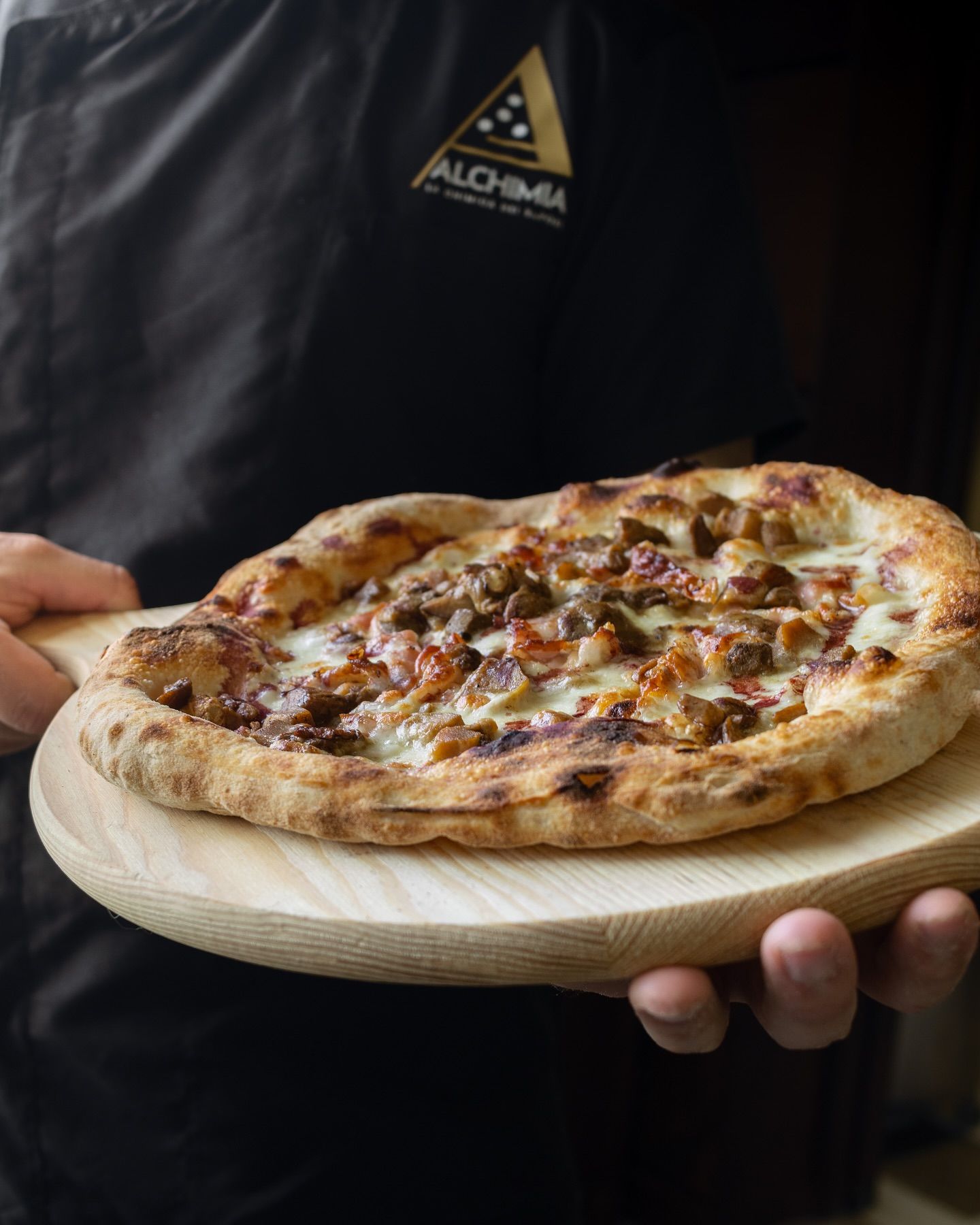 Persona che tiene in mano una pala per pizza in legno con una pizza ricoperta di carne e formaggio, su sfondo scuro.