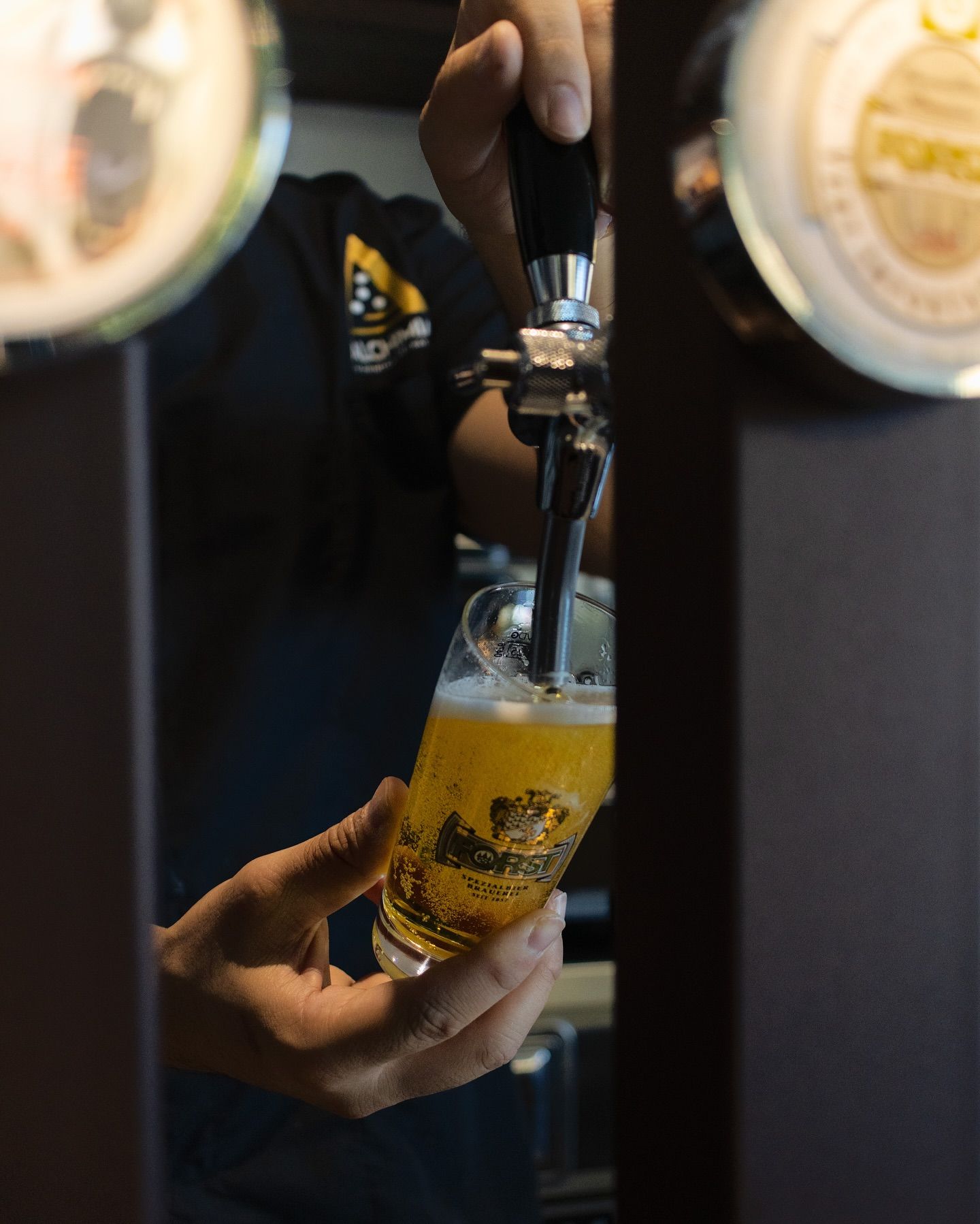 Barista che versa la birra dal rubinetto in un bicchiere da pinta; liquido giallo brillante, colore scuro.