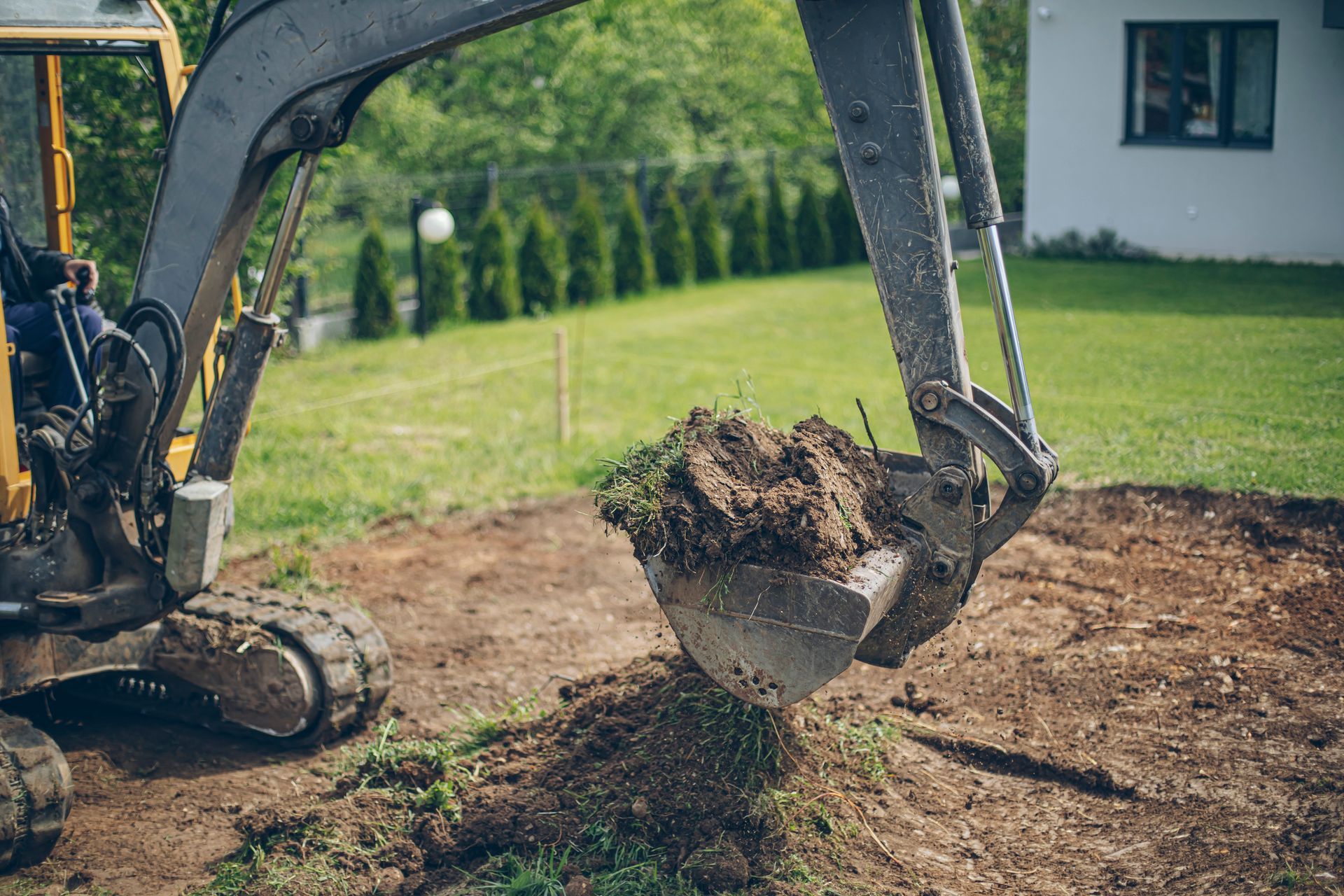 Excavator digging in yard, bucket filled with soil. Green grass, white house in background.