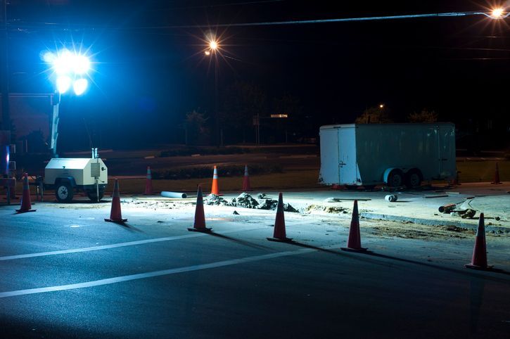Nighttime road construction scene with bright overhead lights, orange cones, and a trailer.
